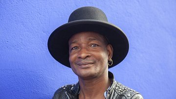Cette image montre un homme souriant, portant un grand chapeau noir qui a une forme de haut de forme. Il est habillé d'une veste en cuir noire ornée de clous, ce qui lui donne un style à la fois décontracté et audacieux. L'arrière-plan est d'un bleu vif, créant un contraste frappant avec ses traits. Son expression est amicale et accueillante, évoquant une personnalité chaleureuse et charismatique.