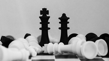 L'image présente deux pièces de jeu d'échecs, un roi noir et un roi blanc, se faisant face au centre. Ils sont placés sur un échiquier, entourés d'autres pièces éparpillées, à la fois noires et blanches. La composition est en noir et blanc, donnant une atmosphère sobre et artistique. Le fond est clair, contrastant avec les pièces sombres, ce qui met en valeur leur forme et détails. L'ensemble évoque une scène de stratégie et de confrontation dans le jeu d'échecs.
