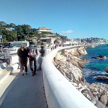 Imaginez-vous sur une promenade longeant la mer. Sur votre droite, vous pouvez entendre le bruit des vagues qui s'écrasent doucement contre les rochers, tandis que l'odeur salée de l'océan flotte dans l'air. Le vent léger caresse votre visage et apporte une sensation de fraîcheur. À votre gauche, des bâtiments se dressent, certains sont des maisons et d'autres des commerces. Vous pouvez sentir l'animation de la ville avec des groupes de personnes qui marchent, certains discutant joyeusement. La promenade est large et bien aménagée, invitant à la flânerie. Au loin, le paysage est ponctué d'arbres et de collines, avec quelques nuages blancs dégradant le bleu du ciel. La lumière du soleil illumine tout le paysage, créant une ambiance chaleureuse. L'ensemble évoque une belle journée au bord de la mer, propice à la détente et au plaisir des sens.