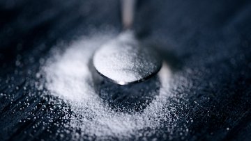 L'image montre une petite cuillère en métal, posée sur une surface sombre. La cuillère contient une fine couche de sucre blanc, qui s'est déversée autour d'elle, créant un léger monticule. La texture du sucre est douce et poudreuse, contrastant avec l'arrière-plan sombre, mettant en valeur la brillance de la cuillère et la pureté du sucre. L'éclairage est doux, accentuant les contours de la cuillère et la délicatesse du sucre dispersé.