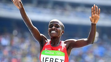 L'image montre une athlète célébrant sa victoire. Elle porte un débardeur rouge avec le nom "JEBET" inscrit dessus, et un logo des Jeux Olympiques sur sa tenue. Ses bras sont levés en signe de triomphe, et elle affiche un large sourire, exprimant la joie et l'excitation de son succès. En arrière-plan, on peut deviner une foule dans un stade, ajoutant à l'atmosphère festive de l'événement. L'éclairage est lumineux, mettant en valeur l'expression de bonheur sur son visage.