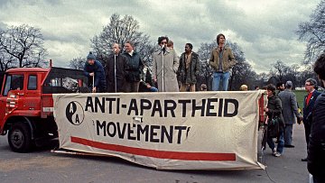 Cette image représente un rassemblement pour le mouvement anti-apartheid. On voit une scène sur un camion, avec plusieurs personnes qui parlent à un microphone. La bannière au premier plan affiche le texte "ANTI-APARTHEID MOVEMENT" en lettres majuscules, souligné par une bande rouge. L'environnement est urbain, avec des arbres dénudés en arrière-plan et un ciel nuageux, ce qui crée une atmosphère sérieuse et engagée. Les participants semblent passionnés, témoignant de l'importance de la lutte pour l'égalité et les droits civiques.