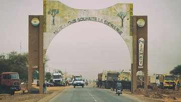 L'image montre une route qui traverse une grande arche d'entrée, symbolisant l'arrivée à Niamey, la capitale du Niger. L'arche est décorée avec des motifs végétaux et des inscriptions en français qui souhaitent la bienvenue. De chaque côté de la route, on peut voir des camions et des véhicules qui circulent, ainsi que des arbres et de la terre poussiéreuse. L'atmosphère est légèrement brumeuse, accentuant le caractère désertique de la région. L'ensemble donne une impression dynamique de la vie et du commerce à l'entrée de la ville.