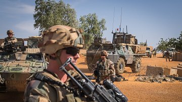 L'image montre un paysage désertique, typique d'une zone militaire. Au premier plan, un soldat est en position, tenant un fusil. Il porte un casque et des lunettes de soleil, ce qui lui confère une allure déterminée. En arrière-plan, on aperçoit d'autres soldats et un véhicule blindé, avec des éléments de terrain militaire, comme des barbelés et des équipements. Le ciel est dégagé, et la lumière du soleil éclaire la scène, soulignant l'environnement aride et la présence militaire. L'atmosphère respire la vigilance et la préparation.