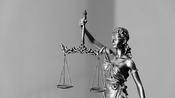 L'image représente une statue de la justice, souvent symbolisée par une femme. Elle est drapée dans une robe et tient une balance dans une main, symbolisant l'équilibre et l'équité. La statue est également dotée d'un bandeau sur les yeux, représentant l'impartialité dans le jugement. L'ensemble de la sculpture est réalisé dans des tons métalliques, et le fond est flou et neutre, mettant en valeur la figure centrale.
