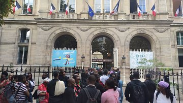 L'image montre un bâtiment imposant avec une architecture classique, visible grâce à ses grandes fenêtres et ses arches. Devant ce bâtiment, un groupe de personnes se rassemble, créant une atmosphère animée. Des drapeaux français flottent au-dessus d'eux, témoignant de l'importance de ce lieu. Les visiteurs semblent variés en termes d'âge et de style vestimentaire, ce qui reflète une diversité culturelle. À l'arrière-plan, des affiches colorées annoncent un événement, ajoutant une touche festive à la scène. L'ensemble dégage une ambiance d'attente et d'excitation.