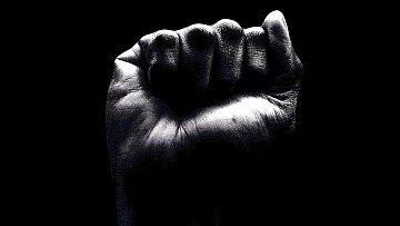 L'image représente un poing fermé, vu de près sur un fond noir. La main est d'une teinte sombre, avec des détails visibles sur la peau, comme les lignes des empreintes digitales et les plis. Le poing, symbolisant souvent la force ou la résistance, est levé de manière à exprimer la détermination. Les contours de la main sont nets, mettant en avant la texture et la forme. L'arrière-plan noir accentue la présence du poing, créant un contraste fort et saisissant.