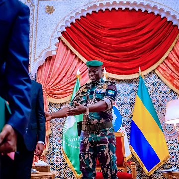 The image shows a scene set in an ornate room. A military officer, dressed in camouflage with a green beret, is smiling and appears to be interacting with another individual. The background features decorative elements, including curtains and flags, likely representing Gabon or an official entity. There are also books and documents in the hands of a person standing to the left, indicating a formal context, possibly a meeting or ceremony. The overall atmosphere seems official and ceremonial.