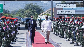 L'image montre un moment protocolaire à un poste frontalier. Deux hommes marchent sur un tapis rouge. À gauche, un homme en costume noir et masque, et à droite, un homme en chemise blanche. De chaque côté du tapis, des soldats en uniforme militaire attendent, formant une haie d'honneur. Les soldats portent des bérets rouges et des masques. En arrière-plan, on peut voir des panneaux indiquant la bienvenue au Rwanda et un poste frontière. L'ambiance est solennelle et cérémonielle.