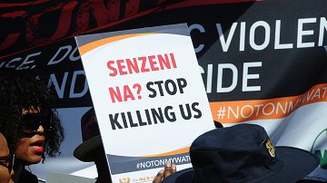 L'image montre un groupe de manifestants rassemblés pour une cause importante. Au premier plan, une personne tient une pancarte avec le slogan « SENZENI NA? STOP KILLING US ». Ce message manifeste un appel urgent à mettre fin à la violence et aux meurtres. En arrière-plan, il y a d'autres bannières et slogans, suggérant une atmosphère de protestation. Les personnes présentes portent des vêtements variés, y compris des chapeaux, et semblent engagées dans la cause qu'elles défendent. L'ambiance générale est celle de la solidarité et de la lutte pour la justice.
