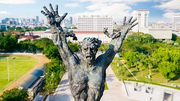 Cette image montre une statue impressionnante d'un homme, représenté dans une pose expressive. Il lève les bras vers le ciel, illustrant un sentiment intense, qui pourrait être celui de la lutte ou de l'angoisse. Son visage est déformé par une expression de détresse, accentuant une impression dramatique. La statue est en bronze, avec une texture riche et des détails finement sculptés qui soulignent les muscles et les traits faciaux. En arrière-plan, on aperçoit une ville moderne avec des bâtiments élevés et des espaces verts, créant un contraste entre la puissance de la sculpture et l'urbanisme environnant. Le ciel est dégagé, ajoutant une luminosité à la scène.