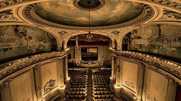 L'image représente l'intérieur d'un ancien théâtre, avec une architecture classique. Le plafond est orné de fresques délicates, tandis que les murs affichent des signes d'usure et de dégradation, témoignant du temps passé. Au centre, un grand miroir est encadré par un drapé rouge, ajoutant une touche de luxe. Devant lui, des rangées de sièges, maintenant poussiéreux, indiquent l'endroit où se tenaient les spectateurs. Des lumières tamisées créent une ambiance mélancolique, et des graffitis sur les murs rappellent l'abandon de ce lieu autrefois vivant. L'ensemble dégage une atmosphère nostalgique, marquée par la beauté d'antan et le passage du temps.