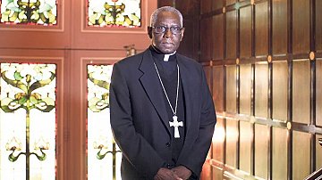 Dans cette image, nous voyons un homme debout dans un environnement qui semble être une pièce élégante, probablement une église ou un bureau religieux. Il porte une tenue noire, typique des membres du clergé, avec une soutane et un col ecclésiastique. Autour de son cou, une grande croix pend, symbolisant son rôle religieux. Le fond est composé de murs en bois foncé et de vitraux colorés, ce qui crée une atmosphère solennelle et respectueuse. Les motifs des vitraux laissent transparaître une lumière douce, ajoutant une dimension spirituelle à l'ensemble de la scène. L'homme a une expression sérieuse, se tenant droit et digne, ce qui évoque l'autorité et la sagesse.