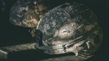 L'image montre deux casques militaires posés sur une surface sombre. Les casques sont de couleur camouflage, avec un motif de nuances grises et vertes. On peut voir des marques d'usure et des imperfections sur la surface, ce qui indique qu'ils ont été utilisés. La lumière frappe légèrement un des casques, mettant en évidence des textures et des détails, tandis que l'autre est dans l'ombre. L'ambiance générale est sobre et évoque un environnement militaire.