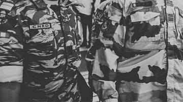 L'image montre des membres de forces armées ou de sécurité, vêtus d'uniformes. À gauche, un soldat porte un uniforme camouflage aux motifs sombres, avec des insignes sur sa poitrine. À droite, un autre soldat est vêtu d'un uniforme de camouflage plus clair, également décoré d'insignes. Les deux uniformes affichent un sens de discipline et d'autorité. L'environnement autour d'eux semble être animé, suggérant une situation dynamique ou un rassemblement. Le contraste entre les motifs des uniformes souligne leur fonction militaire.