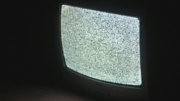 L'image présente un téléviseur ancien, dont l'écran affiche un bruit visuel, souvent appelé "neige", constitué de points blancs et noirs qui scintillent. L'appareil est placé sur le sol, et l'éclairage ambiant est faible, ce qui met en avant le contraste de l'écran lumineux par rapport à l'obscurité environnante. Le téléviseur a des bords arrondis et un design rétro, typique des modèles d'une autre époque. La scène dégage une atmosphère nostalgique et un sentiment d'attente.