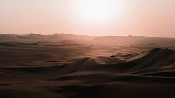 Imaginez une vaste étendue de sable, où les dunes s'étirent à perte de vue, leurs formes ondulantes créant des ombres douces sur le sol. Leurs contours se dessinent en vagues délicates, semblant danser sous les rayons d'un soleil flamboyant qui se lève à l'horizon, infusant le paysage d'une lumière chaude et dorée. Le silence règne, ponctué seulement par le léger murmure du vent qui balaie le désert, et une sensation de calme profonde imprègne l'air. Au loin, on devine une silhouette, peut-être un véhicule, qui semble minuscule face à l'immensité de ce panorama naturel. L'ensemble évoque une beauté brute, une tranquillité presque contemplative, comme si le temps s'était arrêté.