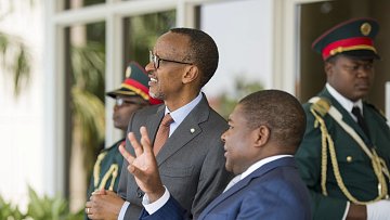 Sur cette image, nous voyons deux hommes en costume dialoguant dans un cadre officiel. L'homme à gauche, avec des lunettes et un sourire, semble être en train d'écouter attentivement. À sa droite, un homme aux cheveux courts gesticule avec les mains, soulignant un point dans la conversation. En arrière-plan, deux gardes en uniforme militaire se tiennent, assurant une ambiance de sécurité. On peut également percevoir des éléments de verdure, suggérant un environnement extérieur agréable.