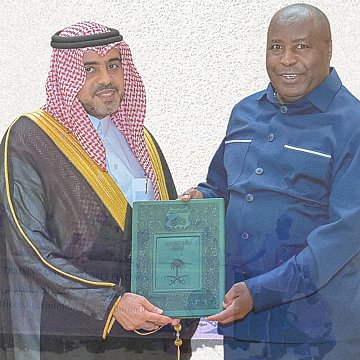 The image depicts a formal ceremony where two men are exchanging a green plaque or certificate. One man is dressed in traditional Saudi attire, including a thobe and a headscarf, while the other is wearing a blue suit. In the background, there is an airplane flying and a group of people holding a banner, possibly in connection to a pilgrimage or religious gathering. The overall tone suggests a celebration or accomplishment related to travel or cooperation, likely involving Saudi Arabia.