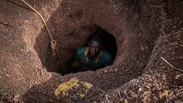 Dans cette image, nous voyons un homme qui se tient au fond d'un trou creusé dans le sol. Le trou a une forme ronde, et les parois sont en terre brune, légèrement humides et irrégulières. L'homme, vêtu d'un maillot vert avec un col jaune, regarde vers le haut avec une expression sérieuse. Son visage est en partie dans l'ombre, mais son regard est visible. Autour du trou, on peut apercevoir des gravats et des brindilles qui ajoutent un aspect naturel et brut à l'environnement. La scène évoque un sentiment d'isolement et de travail ardu.