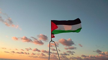 The image depicts a flag flying on a boat against a backdrop of a colorful sky during sunset. The flag features three horizontal stripes: black on the top, white in the middle, and green on the bottom, with a red triangle on the left side. The ocean can be seen below, and there are clouds in the sky that are illuminated by the setting sun, creating a serene atmosphere.