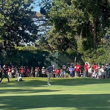 Imaginez un vaste terrain de golf, baigné par la lumière du soleil. Au premier plan, deux joueurs se concentrent sur le putting green, faisant preuve de délicatesse et de précision. L'un d'eux est en position de putt, tandis que l'autre observe attentivement. À l'arrière-plan, des spectateurs en tenue décontractée, vêtus de couleurs variées, attendent avec excitation, certains filmant ou prenant des photos. Autour du green, de grands arbres fournissent de l'ombre, ajoutant à la beauté de l'environnement. À droite, on aperçoit un bâtiment avec des balcons, où d'autres personnes sont rassemblées pour profiter du spectacle. L'atmosphère est vivante, remplie de murmures, de rires et de l'anticipation d'un moment décisif dans le jeu.