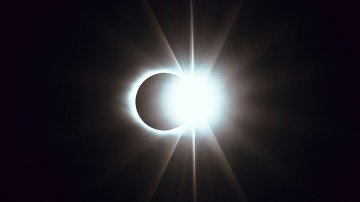 L'image représente un phénomène astronomique spectaculaire : une éclipse solaire. Au centre, un cercle éclatant de lumière intense, représentant le soleil, est partiellement recouvert par la silhouette sombre de la lune. Cette superposition crée un effet visuel époustouflant, où des rays lumineux jaillissent autour du bord de la lune, formant une sorte de couronne brillante. Le contraste entre la lumière vive et l'ombre de la lune évoque une atmosphère immersive, presque mystique, qui attire l'attention et suscite l'émerveillement face à la beauté de l'univers.