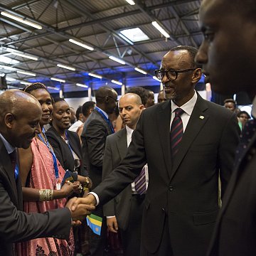 Dans cette image, nous voyons un groupe de personnes rassemblées dans un espace intérieur, probablement lors d'un événement officiel ou d'une conférence. Parmi eux se trouve un homme en costume noir avec une cravate, qui semble être un dirigeant ou une personne importante. Il tend la main pour saluer un autre homme, également en costume. Les autres personnes présentes, principalement habillées formellement, regardent avec intérêt. L'atmosphère est conviviale et respectueuse, avec des sourires et des échanges amicaux. Les murs sont décorés de manière simple, et un éclairage doux illumine la scène.
