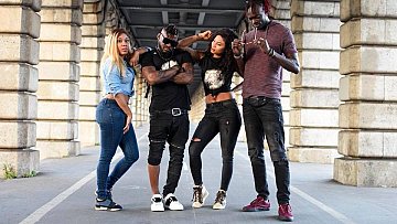 L'image présente un groupe de quatre personnes se tenant sous un pont, avec des colonnes en pierre sur les côtés. À gauche, une femme porte une chemise en denim et un jeans serré, affichant une posture confiante. À côté d'elle, un homme avec un look musclé, portant un tee-shirt noir, se distingue par ses lunettes de soleil et sa barbe. Il est entouré de deux autres femmes. L'une d'elles a les cheveux longs et noirs, vêtue d'un haut noir et d'un jeans. Elle se tient légèrement penchée, adoptant une attitude détendue. La dernière personne est un homme avec des cheveux colorés, aussi habillé de manière décontractée, tenant un appareil dans ses mains. L'environnement donne une ambiance urbaine, renforcée par le béton et les ombres créées par la structure du pont.