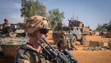 The image depicts a military scene in a desert environment. In the foreground, a soldier wearing a helmet and sunglasses is holding an assault rifle, looking serious and vigilant. Behind him, there are other soldiers and military vehicles, including an armored personnel carrier. The background shows a clear blue sky with some greenery and sand, indicating a remote and arid location. The overall atmosphere suggests a sense of readiness and military operations.