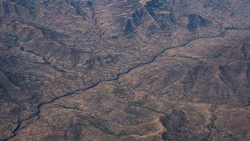 Cette image montre un paysage désertique vu du ciel. On peut observer de vastes étendues de terrain aride, striées par des vallées et des collines aux formes irrégulières. Au centre, un cours d'eau serpente à travers ce paysage, créant un contraste avec les zones sèches environnantes. Les couleurs dominantes sont des teintes de marron et de beige, évoquant un sol rocailleux et dénudé, tandis que la rivière apporte une touche de reflets plus sombres, suggérant des zones humides dans ce milieu. L'ensemble dégage une impression de vastitude et de sérénité, avec une nature sauvage encore peu altérée par l'homme.