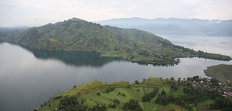 The image showcases a breathtaking landscape featuring lush green hills and a large lake. The hills are gently sloped and dotted with patches of vegetation, indicating a fertile area. In the foreground, there are scattered houses or structures, while the calm waters of the lake reflect the surrounding scenery. In the background, more mountainous terrain rises, adding depth to the view under a slightly overcast sky. The overall atmosphere is serene and picturesque, highlighting the natural beauty of the region.