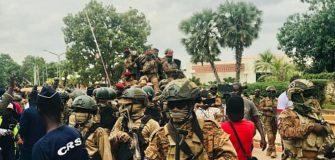 L'image montre une scène animée en plein air, probablement lors d'un événement public ou d'une manifestation. Au premier plan, plusieurs soldats sont regroupés, portant des uniformes militaires et des casques. Ils sont armés et semblent vigilants. L'un d'eux est sur un véhicule militaire, tandis que d'autres avancent sur la route. En arrière-plan, des arbres et des bâtiments peuvent être aperçus, et une foule de citoyens se trouve à proximité, certains portant des vêtements colorés. L'atmosphère peut sembler tendue, surtout en raison de la présence militaire. Le ciel est nuageux, ajoutant une certaine gravité à la scène.