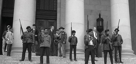 L'image montre un groupe de personnes sur les marches d'un bâtiment imposant, probablement une institution gouvernementale. Au premier plan, plusieurs jeunes hommes se tiennent debout, portant des vestes sombres et des armes à feu, avec des expressions déterminées sur leurs visages. Ils sont entourés d'autres individus, certains vêtus de manière formelle, comme des costumes et des chapeaux. Tous semblent engagés dans une sorte de manifestation ou de rassemblement, émettant un sentiment de tension et de détermination. L'architecture du bâtiment est majestueuse, avec de grandes colonnes en arrière-plan. L'atmosphère générale de l'image évoque une période de lutte pour les droits civiques.