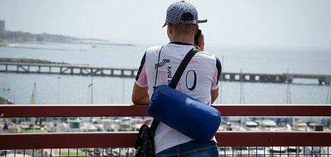 Dans cette image, un homme se tient de dos, regardant au loin vers la mer. Il est vêtu d'un t-shirt blanc avec des accents colorés et porte une casquette à motifs. Sur son épaule, il a un petit sac bleu. L'arrière-plan montre un port avec des bateaux amarrés et une jetée qui s'étend dans l'eau, créant une atmosphère tranquille. Le ciel est clair, suggérant une journée ensoleillée. L'homme semble concentré sur ce qu'il observe, profitant de la vue sur l'horizon maritime.